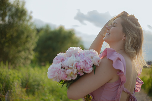 Happy women florist, flower farmer with bouquet of soft pink peonies laughs on summer day in nature.