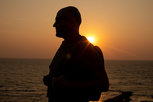Portrait of traveler in sunglasses walking with backpack near sea on seashore at sunset, standing against seascape.