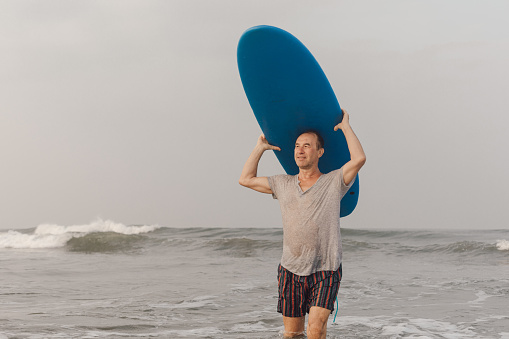 Sporty middle aged surfer coming out of water with blue surfboard on head and looking towards shore standing in sea.