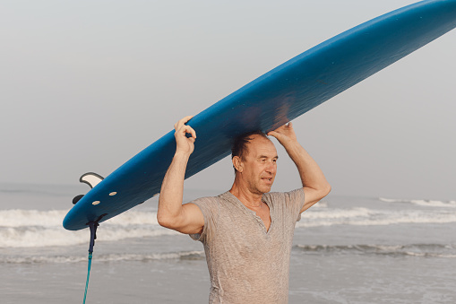 Middle aged athlete carrying blue surfboard on head and holding surfing equipment with hands coming out of waving sea.