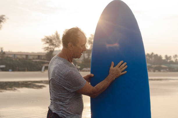 istock Middle aged athlete standing on sandy beach at sunset and touching blue surfboard, checking equipment before activity. 1833614019