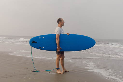 Mature athlete keeping blue surfboard under armpit standing on wet sandy shore in front of sea and looking at seascape.