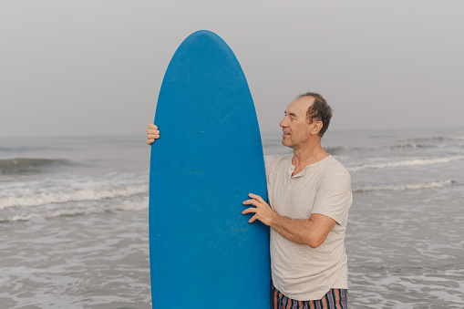 Middle aged surfer holding blue surfboard close to body standing in sea against seascape and looking towards board.