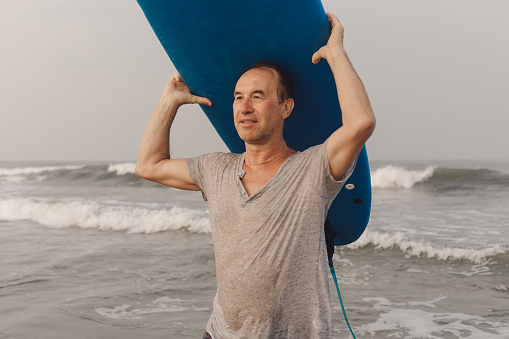 Calm surfer carrying blue surfboard on head, holding equipment with hands and looking ahead on background of seascape.