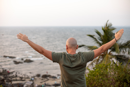 Rear view of man standing against seascape with arms wide open, looking towards waving sea, spending time near beach.