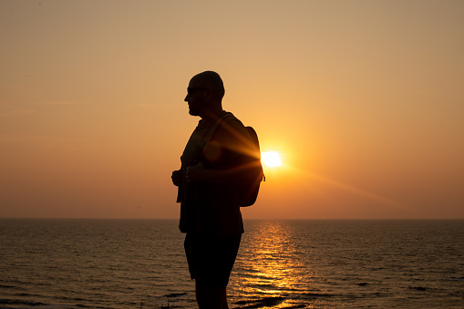 Silhouette of bald vacationer in sunglasses with backpack standing sideways against seascape on seaside at sunset.