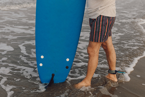 Legs of man with surfing leash attached to ankle standing in water on sandy seashore with surfboard and entering sea.