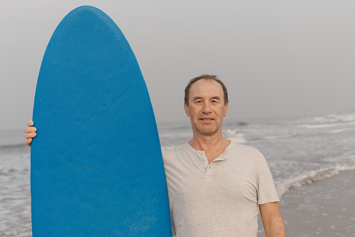 Portrait of middle aged surfer standing on waving seaside against seascape, holding blue surfboard and looking ahead.