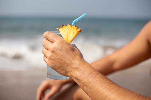 Hand of man holding glass of drink with blue straw decorated with piece of pineapple relaxing on sunny shore near sea.