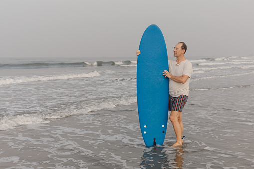 Relaxed mature surfer standing in waving sea on seashore and holding blue surfboard close on background of seascape.