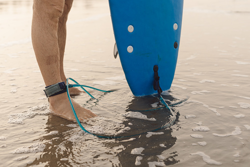 Legs of man standing on sandy seaside with leash attached to ankle, holding surfboard. Athlete preparing for surfing.