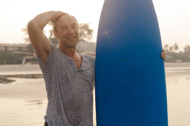 istock Happy mature man with surfboard standing on sandy beach and touching bald head while looking at distance on sunset. 1833651326