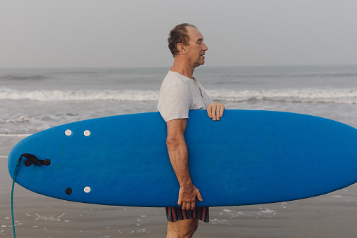 Confident mature surfer standing sideways on sandy seashore and holding blue surfboard under armpit against seascape.