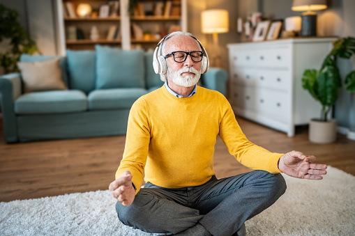 Elderly man, pensioner, sitting on floor practicing guided meditation at home, relaxing body and mind concept