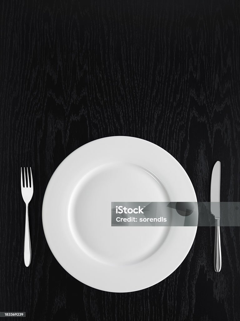 A white plate, a knife and a fork on a black table. Directly above view of table setting on a black table Gardening Fork Stock Photo