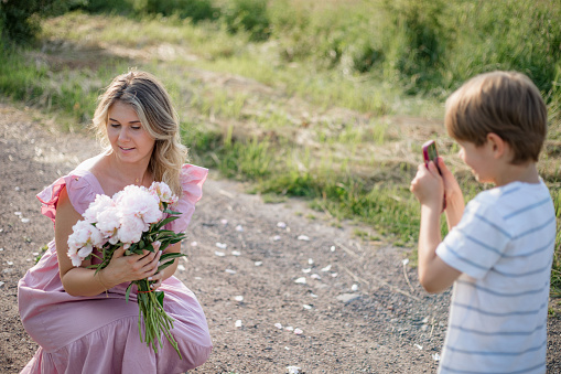 Preschooler son photographs his happy mother with bouquet of peonies on summer day in countryside.