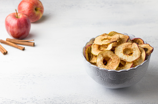 Homemade dried apples with cinnamon, apple chips in a bowl with cinnamon sticks on a light gray background. Delicious healthy snack.