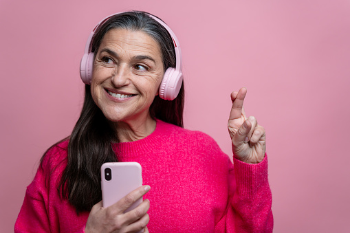 Portrait of happy senior woman in pink sweater listening to music with headphones and mobile phone crossing fingers