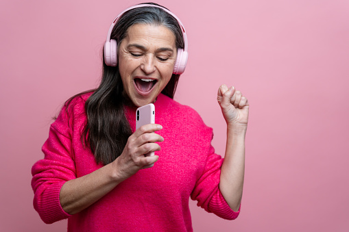 Portrait of a happy senior woman listening to music with headphones on a pink background