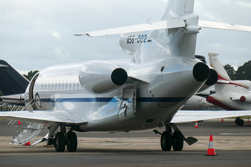 A Royal Australian Air Force Dassault Falcon 7X plane, registration A56-002, used for VIP transport of the Prime Minister and others in government and military, on the tarmac at Sydney Kingsford-Smith Airport.  She arrived as flight EVY42 from Canberra at 4.45pm and will return as the same flight number at 8pm. The cargo door is open. She is parked in an area for corporate jets, some of which are visible in the background. Her fuselage engine is running, causing a heat haze.  This image was taken from behind the steel security fence on Ross Smith Avenue, Mascot on an overcast afternoon on 3 December 2023