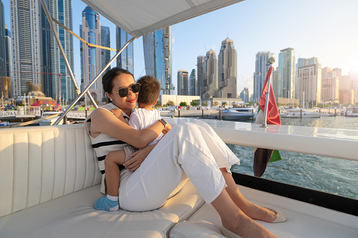 In the enchanting glow of the Dubai Marina sunset, a mother and her toddler share a tender embrace on a boat ride. Their connection is illuminated by the warm hues of the setting sun, creating a heartwarming scene of love and togetherness against the city skyline