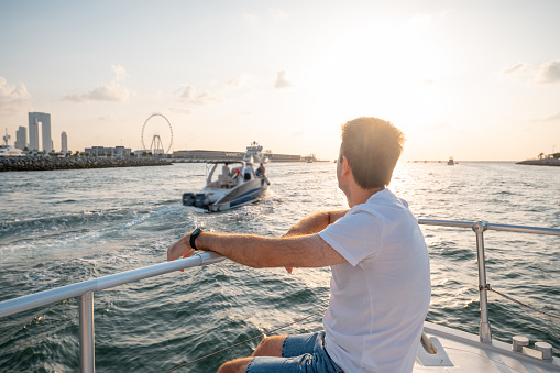 Adult man embraces the splendor of Dubai skyline as he siting on a yacht, captivated by the enchanting sunset over Dubai Marina. This tranquil moment exudes luxury and relaxation