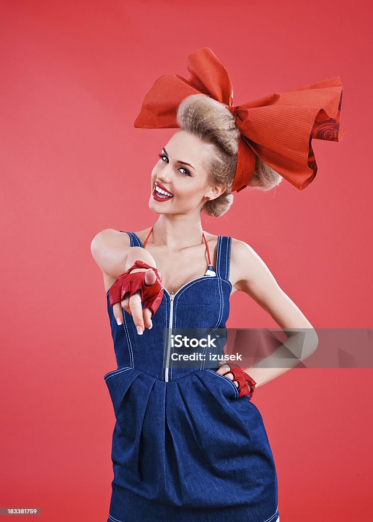 Beautiful girl pointing at camera "Smiling Young blond woman wearing jeans dress, red gloves and big, red hair ribbon pointing at viewer. Standing against red background. Studio shot" 20-24 Years Stock Photo