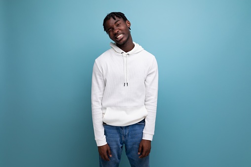 positive african young brunette man in white hoodie with cute smile on blue background.