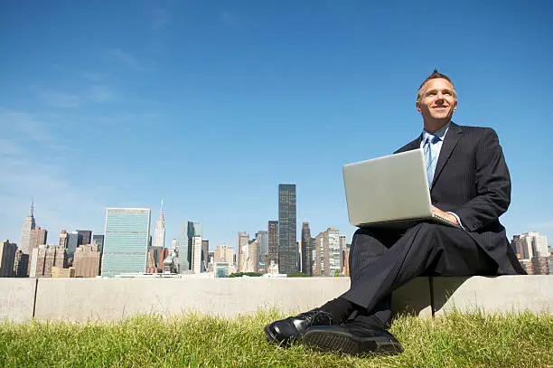 Photo of Smiling Businessman Sitting Using Laptop Outdoors by City Skyline