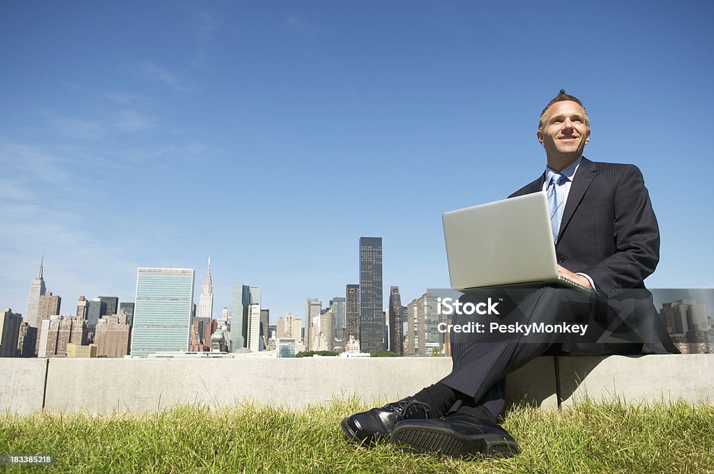 Uśmiechająca się Biznesmen siedzi przy użyciu laptopa na zewnątrz, z widokiem na panoramę miasta - Zbiór zdjęć royalty-free (Laptop)