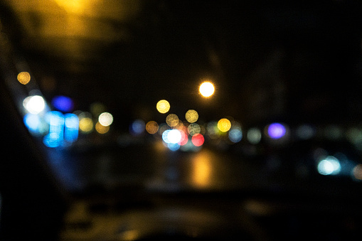 Driver's point of view through a dark night car windshield looking ahead at defocused traffic, vehicle and city lights of various bright colors.