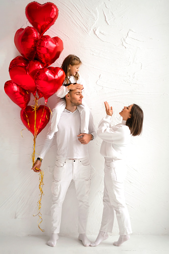 A young couple with a baby and red heart-shaped balloons on Valentine's day. Daughter sits on her father's neck, mother blows them a kiss