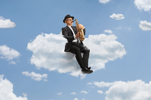 Mature male artist playing a saxophone seated on a cloud up in the sky
