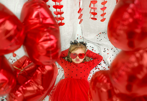A girl in a red festive dress and glasses lies surrounded by foil balloons , folded her lips in a kiss