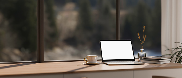 A white-screen laptop computer mockup, a coffee cup, book, and copy space on a wooden tabletop against the glass window in a modern room. Workspace concept. 3d render, 3d illustration
