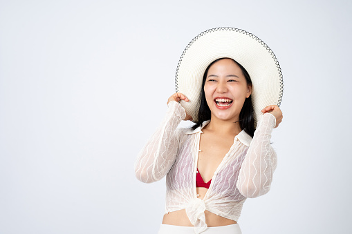 A beautiful Asian woman in summer clothes and a white straw hat is posing cute, showing happy facial expression, isolated white background with a copy space. summer vacation, holiday trip, beach