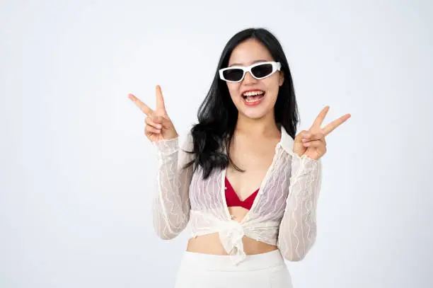A cheerful and positive Asian woman in summer clothes and sunglasses is showing two-fingers peace hand sign and smiling at the camera, isolated white background. Summer vacation, beach holiday trip