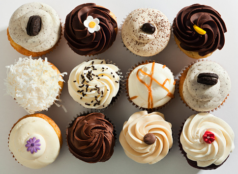 Subject: Horizontal high angle, directly overhead view of a dozen frosted cupcakes. The cupcakes are against a white background and are decorated with swirls, sprinkles, drizzles and candies—a tempting decadent treat for those who indulge in sweets and love cakes with lots of toppings.