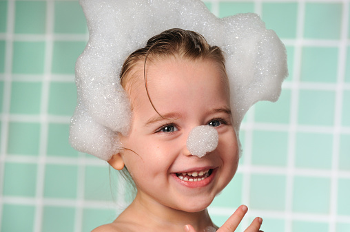 Horizontal image of an adorable young girl who is having fun in her bubble bath. She is piling the bubbles on top of her head and her nose while giggling. She has a beautiful smile and sparkling eyes. 
