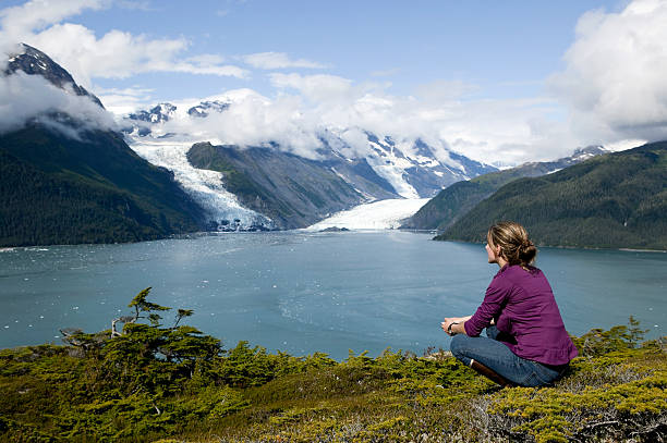 Woman sitting and gazing at water and snowy hills in Alaska one adult woman in prince william sound alaska overlooking columbia glacier chugach mountains photos stock pictures, royalty-free photos & images