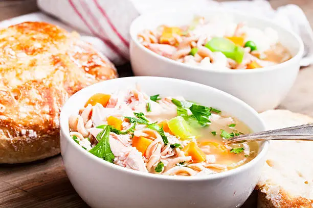 2 bowls filled with healthy chickensoup,bowl filled with healthy chicken soup, made of chicken, whole wheat pasta, and lots of vegetables, on wooden background, with some bread