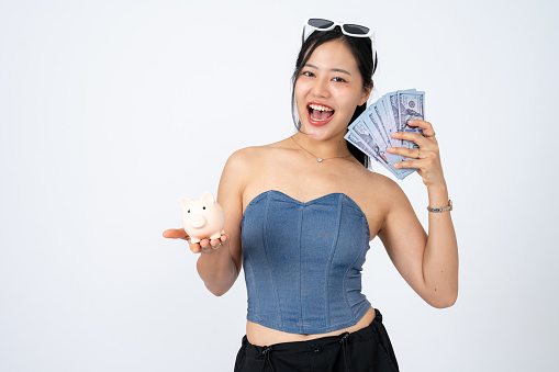 A beautiful and positive Asian woman in trendy clothes is holding a piggy bank and cash on an isolated white background. financial, banking, shopping, saving