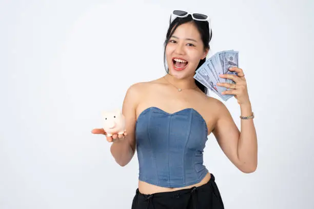 Photo of A beautiful Asian woman is holding a piggy bank and cash on an isolated white background.