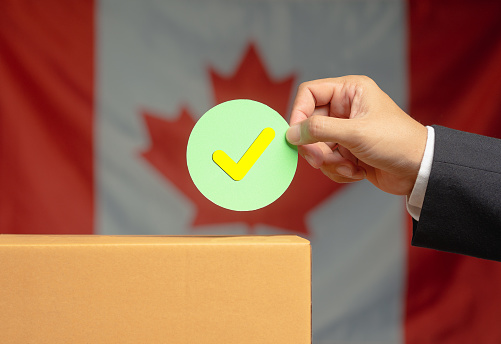 Elections in Canada. Hand holding a check mark symbol overhead the voting box at place election with the Canada flag background. Campaign to exercise the right to vote