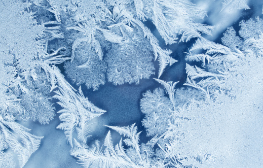 An intricate crystalline pattern of frost  shaped like feathers and moss against a stream on a window.