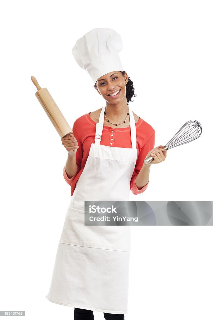 Smiling Young Woman Chef with Apron, Hat, and Kitchen Utensils "Subject: Vertical three-quarter figure portrait of a smiling young black woman wearing a white apron and chefaas hat and holding a rolling pin and whisk, leaning and tilting her head as if almost dancing, happy about cooking. Photographed on a white background." One Woman Only Stock Photo