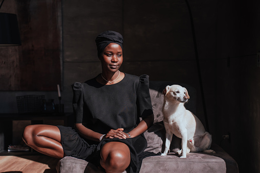 Young African girl in black turban and black traditional clothes sitting on couch with white dog being in pensive mood, relaxing at home on sunny summer day. Companion.