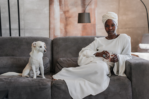 African girl in white turban and traditional dress sitting on couch with  dog relaxing at home looks at camera with confident face.