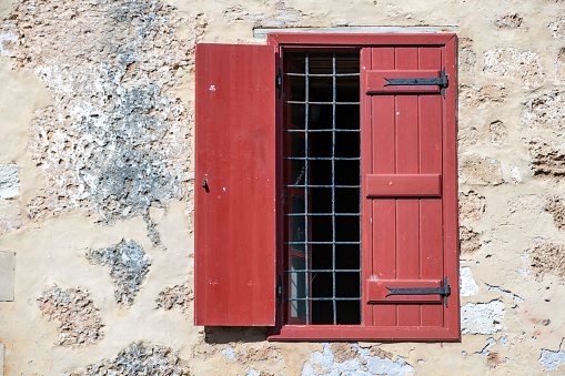 Window with wooden red shutter and metal grid on weathered peeled wall background. House front view, Greek traditional architecture, sunny day.