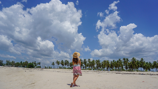 Back view of asian women in a straw hat standing breathe fresh air on the beach, coconut garden and fluffy clouds clear sky, Feeling free, Travel on holiday or summer vacation.